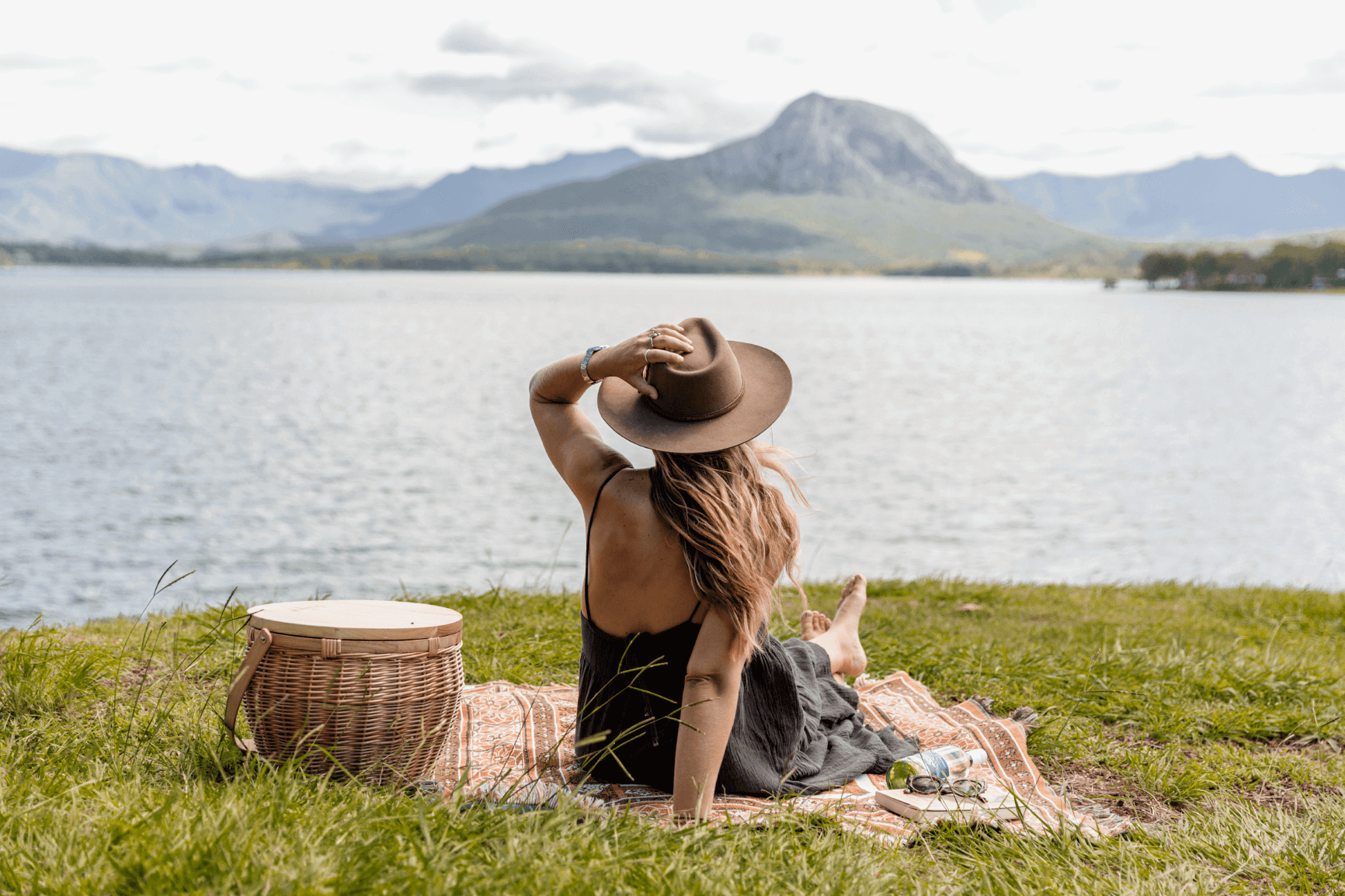 A young woman sits on a picnic rug by Lake Moogerah within the Scenic Rim in Queensland, Australia. She is looking at the mountains across the lake and she holds her hand on her hat to stop it blowing away.