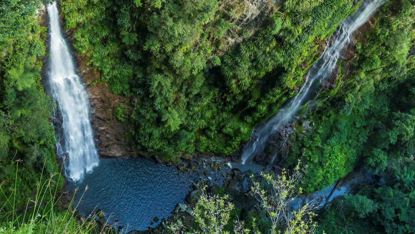 View down sheer rock wall with two waterfalls and pool at bottom.