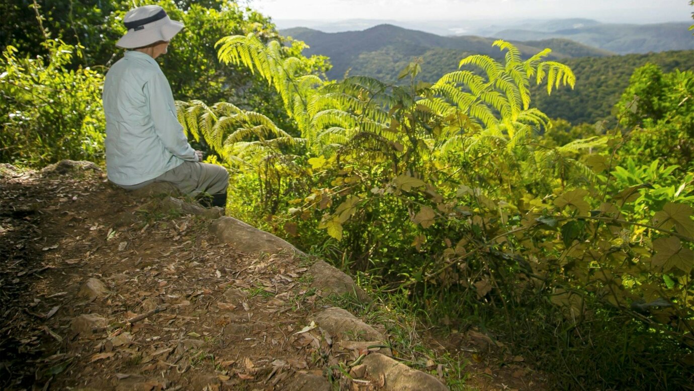 Walker sits on rock ledge looking over wideranging view of mountains in distance.